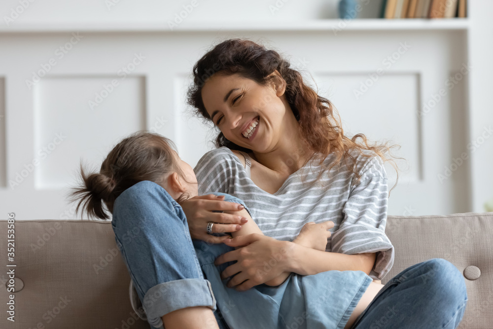 Smiling young mother holding little daughter, sitting on cozy couch in living room, laughing loving mum hugging cute preschool girl, family having fun together, spending leisure time at home
