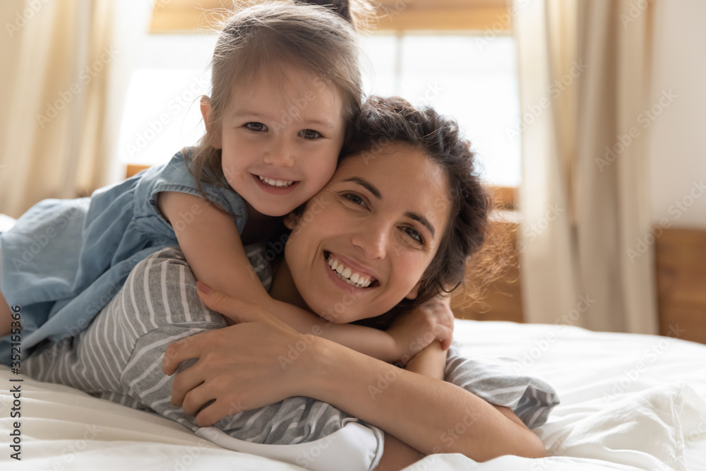 Head shot portrait little girl lying on happy mother back, hugging and cuddling, beautiful young mum and preschool daughter looking at camera, family enjoying leisure time, tender moment in bedroom Head shot portrait little girl lying on happy mother back, hugging and cuddling, beautiful young mum and preschool daughter looking at camera, family enjoying leisure time, tender moment in bedroom