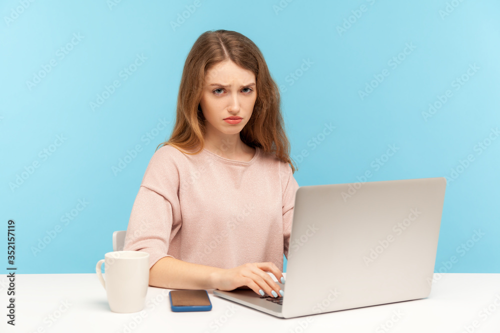 Gloomy unhappy young businesswoman in casual clothes looking at camera with frustrated depressed face, working on laptop at home office, freelance job. indoor studio shot isolated on blue background Gloomy unhappy young businesswoman in casual clothes looking at camera with frustrated depressed face, working on laptop at home office, freelance job. indoor studio shot isolated on blue background