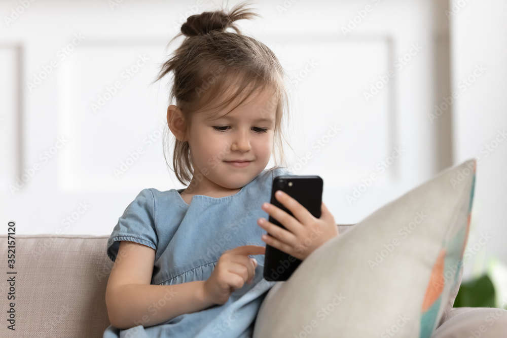 Close up curious cute little girl holding phone, looking at screen, playing mobile device game, watching cartoons online, making video call to grandparents, sitting on cozy couch at home Close up curious cute little girl holding phone, looking at screen, playing mobile device game, watching cartoons online, making video call to grandparents, sitting on cozy couch at home