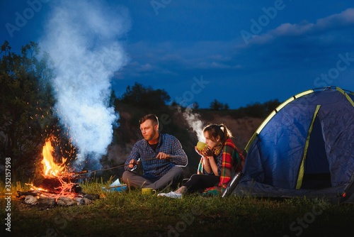 Young couple having a rest at bonfire beside camp and blue tourist tent, drinking tea, enjoying night sky. The concept of active recreation and travel with a tent
