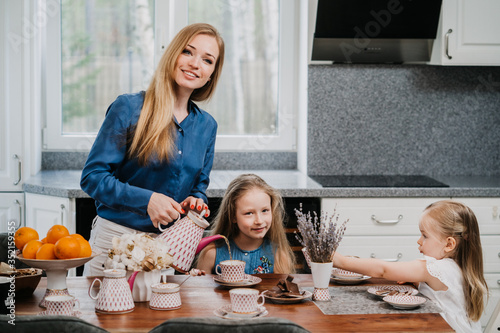 Happy family shot in the kitchen. Young beautiful woman pours a tea for her daughters sitting at the table, enjoying their breakfast. Mother and daughters.