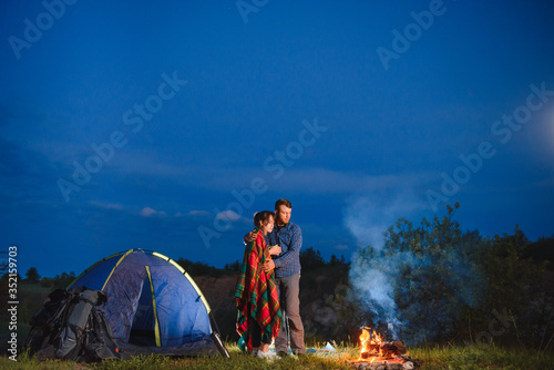 Night camping in the mountains. Happy couple travellers sitting together beside campfire and glowing tourist tent. On background big boulder, forest and night sky.