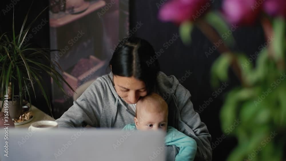 Young mother with a 6 month old baby in her arms works on a laptop while sitting at a table in the kitchen. in slow motion Young mother with a 6 month old baby in her arms works on a laptop while sitting at a table in the kitchen. in slow motion