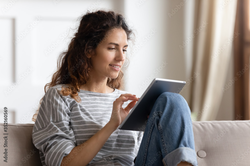 Smiling young woman using computer tablet, sitting on couch, typing on touchscreen, chatting in social network with friends, playing mobile device game or surfing internet, shopping online Smiling young woman using computer tablet, sitting on couch, typing on touchscreen, chatting in social network with friends, playing mobile device game or surfing internet, shopping online