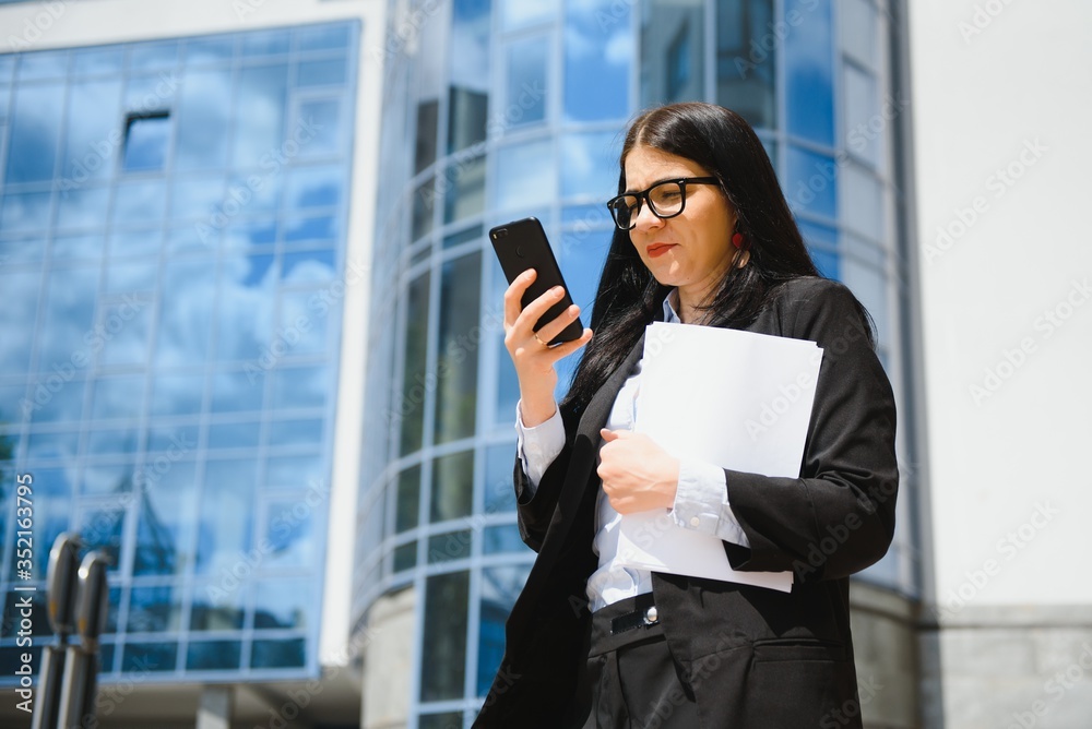 Business Woman With Phone Near Office. Portrait Of Beautiful Smiling Female In Fashion Office Clothes Talking On Phone While Standing Outdoors. Phone Communication. High Quality Image. Business Woman With Phone Near Office. Portrait Of Beautiful Smiling Female In Fashion Office Clothes Talking On Phone While Standing Outdoors. Phone Communication. High Quality Image.