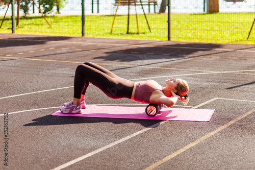 Sporty blond woman training on mat outdoor summer day, using foam roller massager on her upper back for relaxation, stretching spine muscles, doing fascia exercise. Health care, workouts routine