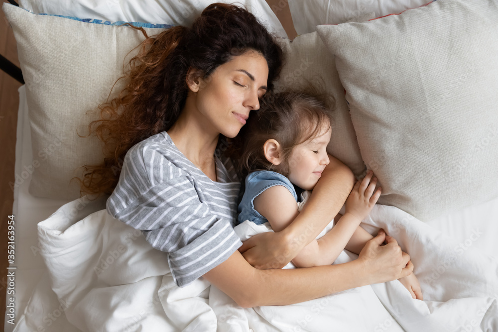 Top view close up peaceful loving mother and little daughter sleeping on soft pillow together, relaxing lying in cozy bed under warm blanket, caring young mum hugging sleepy preschool girl