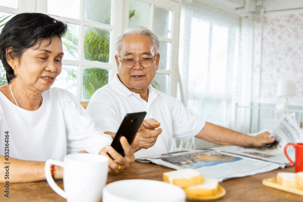 Portrait of an elderly Asian couple following the daily news by reading a newspaper and smartphone while eating breakfast. They enjoy and smiling faces. The activity of retirement age Portrait of an elderly Asian couple following the daily news by reading a newspaper and smartphone while eating breakfast. They enjoy and smiling faces. The activity of retirement age