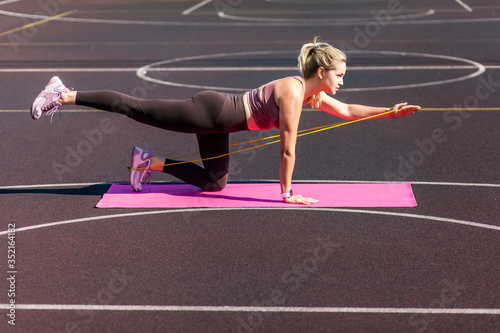 Motivated, concentrated athletic woman doing sports, training on mat outdoor, exercising with rubber expander stripe, using elastic rope for muscle stretching and strength workouts. Health care © khosrork
