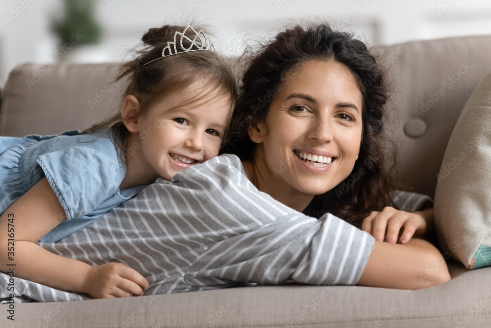 Head shot portrait cute girl wearing princess diadem lying on happy mother back, adorable daughter with mum relaxing on cozy couch together, looking at camera, family enjoying lazy weekend at home Head shot portrait cute girl wearing princess diadem lying on happy mother back, adorable daughter with mum relaxing on cozy couch together, looking at camera, family enjoying lazy weekend at home