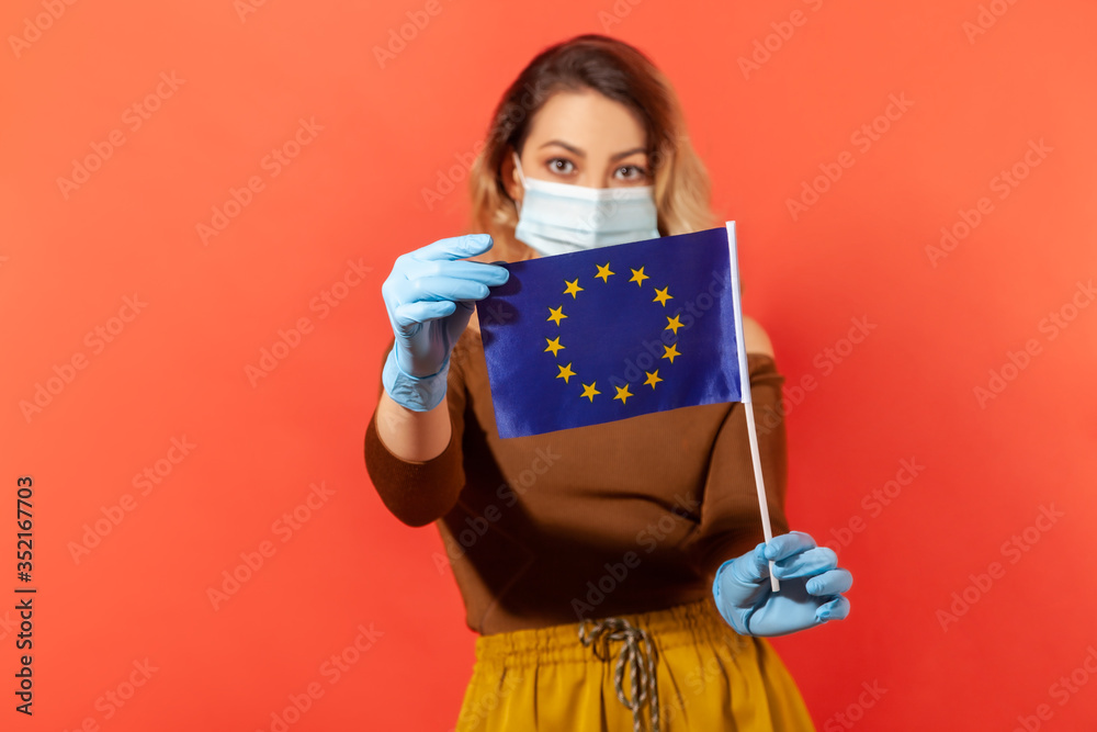 Fototapeta premium Woman in hygienic face mask and gloves showing European Union flag, concept of coronavirus pandemic threat in Europe, closed borders, visa abolition and travel restriction. indoor studio shot isolated