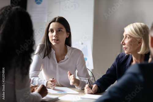 Confident young businesswoman speaking at corporate meeting, female intern sharing startup ideas, discussing project strategy with colleagues at briefing, group business partners negotiation