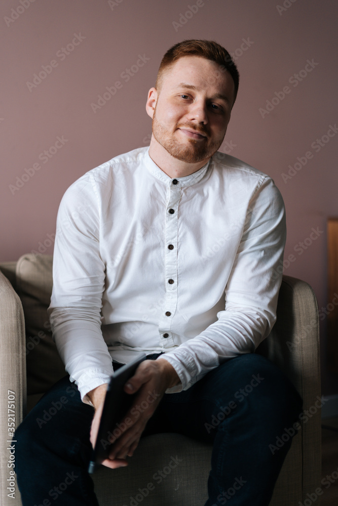 Young photogenic man sitting in white soft armchair posing with digital tablet in hands, looking at camera Young photogenic man sitting in white soft armchair posing with digital tablet in hands, looking at camera