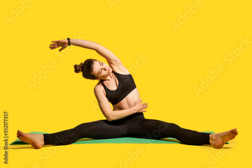 Gymnast girl with hair bun in tight sportswear sitting on mat with spread legs, raising hand bending to side, doing stretching muscle workouts, flexibility exercises. studio shot, isolated on yellow