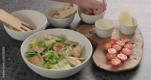 Man hand add cherry tomatoes and chicken on caesar salad in white ceramic bowl on concrete countertop