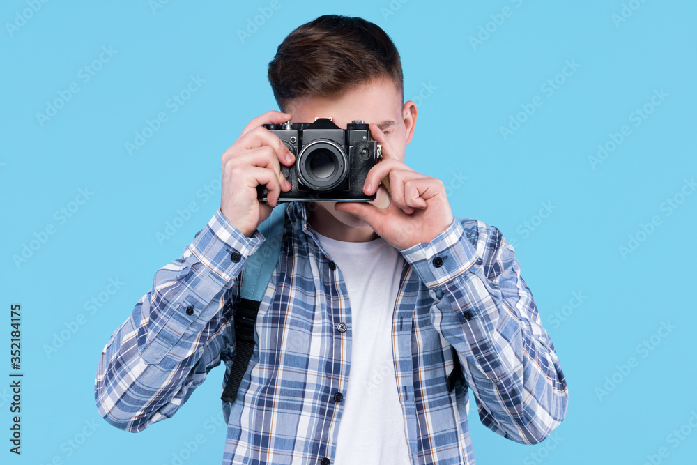 Young man photographer in white t-shirt, checkered shirt, is holding retro photo camera, standing on blue background. Guy student, traveller is making photography. Travel, trip, journey concept. Young man photographer in white t-shirt, checkered shirt, is holding retro photo camera, standing on blue background. Guy student, traveller is making photography. Travel, trip, journey concept.