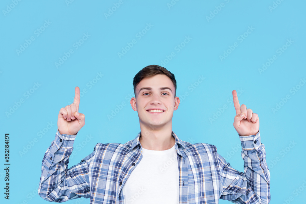 Cheerful smiling guy dressed in white t-shirt, checkered shirt is standing on blue background. Young man is showing direction with hand to up. Male gesturing. Emotional portrait concept. Cheerful smiling guy dressed in white t-shirt, checkered shirt is standing on blue background. Young man is showing direction with hand to up. Male gesturing. Emotional portrait concept.