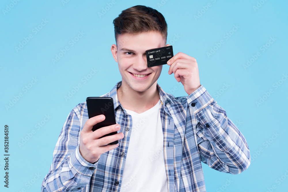 Young smiling man in white t-shirt, checkered shirt is holding black credit card, smartphone, standing on blue background. Guy student is going to pay for purchase cashless. Shopping and sale concept. Young smiling man in white t-shirt, checkered shirt is holding black credit card, smartphone, standing on blue background. Guy student is going to pay for purchase cashless. Shopping and sale concept.