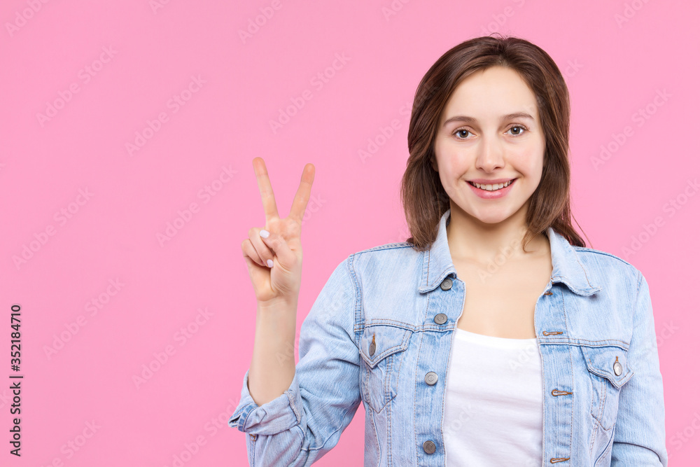 Cheerful smiling girl dressed in white t-shirt, denim jacket on pink background. Young woman is showing with fingers peace sign, number two. Female gesturing. Emotional portrait concept. Cheerful smiling girl dressed in white t-shirt, denim jacket on pink background. Young woman is showing with fingers peace sign, number two. Female gesturing. Emotional portrait concept.