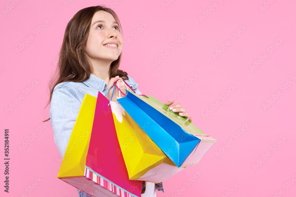 Young woman in white t-shirt, denim jacket is holding colorful paper bags, packages, packets. Smiling girl is rejoicing in purchases, standing on pink background. Shopping and sale concept.