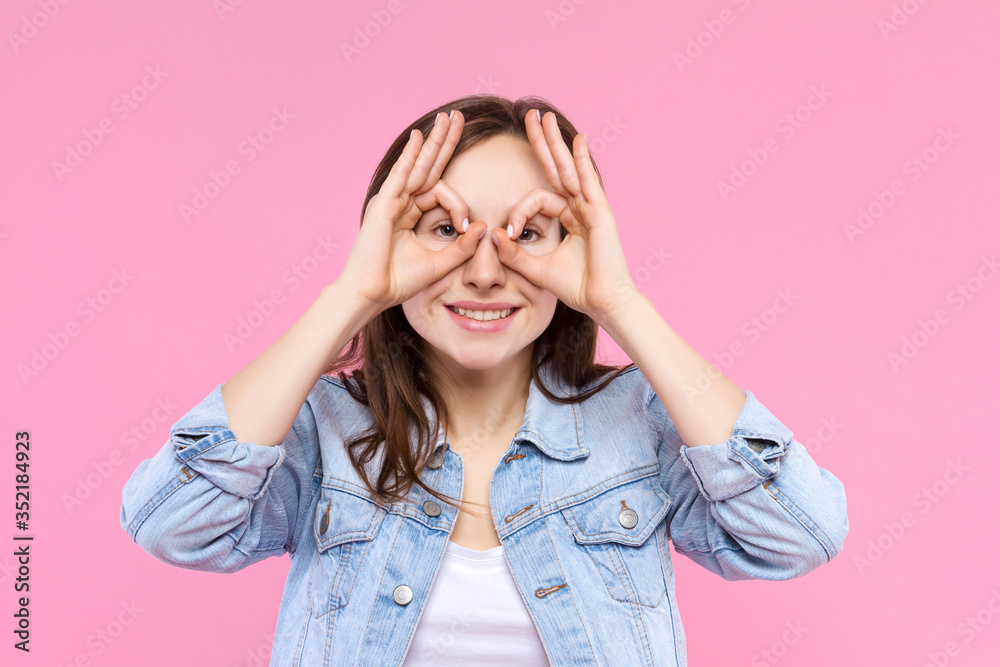 Cheerful smiling girl dressed in white t-shirt, denim jacket on pink background. Young woman is grimacing, showing glasses, mask with hands. Female gesturing. Emotional portrait concept. Cheerful smiling girl dressed in white t-shirt, denim jacket on pink background. Young woman is grimacing, showing glasses, mask with hands. Female gesturing. Emotional portrait concept.