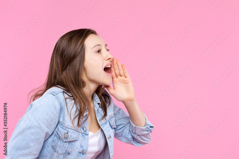 Cheerful smiling girl dressed in white t-shirt, denim jacket on pink background. Young woman is screaming, shouting into hands folded in loudspeaker. Female gesturing. Emotional portrait concept. Cheerful smiling girl dressed in white t-shirt, denim jacket on pink background. Young woman is screaming, shouting into hands folded in loudspeaker. Female gesturing. Emotional portrait concept.