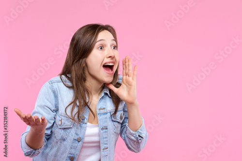 Cheerful smiling girl dressed in white t-shirt, denim jacket on pink background. Young woman is screaming, shouting into hands folded in loudspeaker. Female gesturing. Emotional portrait concept.