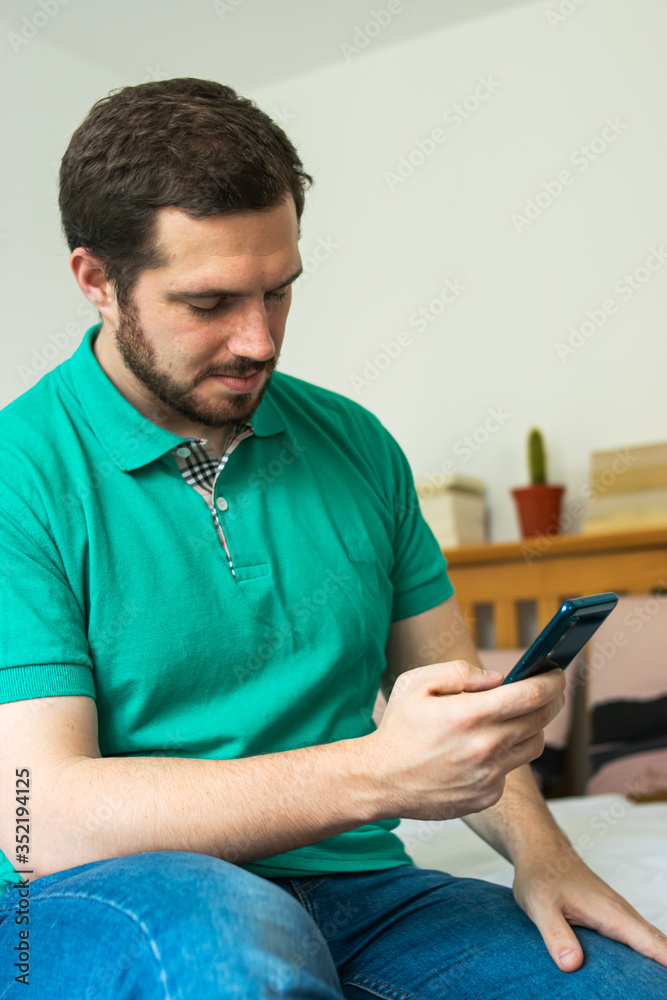 Photo of a young and attractive man with beard checking his phone sitting in the bed with a relax expression