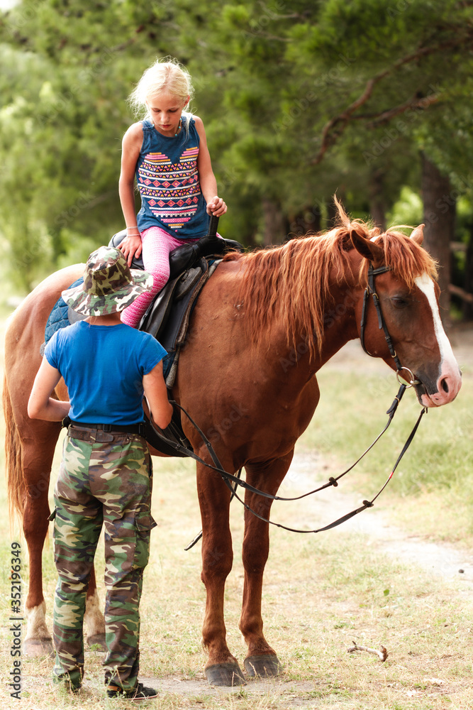 Obraz premium The instructor helps the girl to saddle a brown horse in the forest. Children's equestrian camp. Summer sports camp for children. A nice girl is learning to ride a horse.