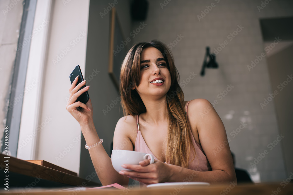 Young positive woman with smartphone and cup of coffee at home