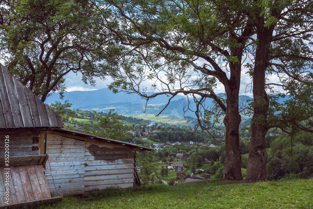 An old wooden house is located on a hill. The village and mountains are further away.