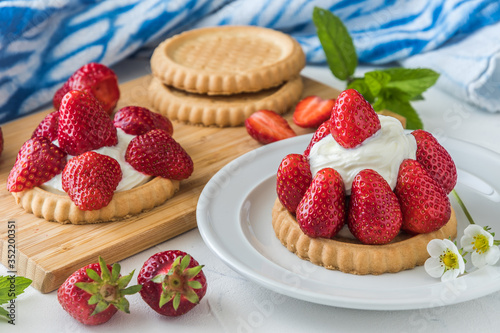 Strawberry tartlet on white plate  decorated with blossoms and mint leaves  in the background wooden board with tartlets and strawberries and blue and white patterned cloth.