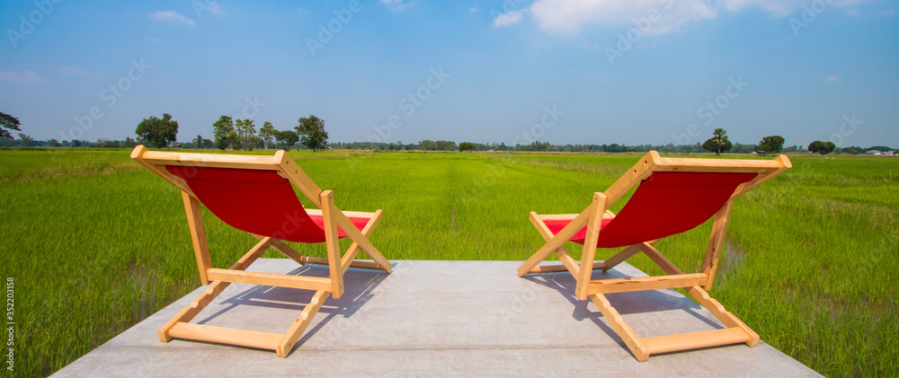 The back view of Two wooden folding chairs For sitting on the lost area The backseat is red and the seat is empty. The background is lush green rice fields and the sky is beautiful, with some clouds.