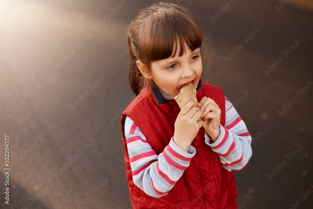 Close up portrait of funny playful little kid being outside, having walk, looking ahead, holding food in hands, eating ice cream, wearing casual clothes, enjoying vacation. Childhood concept. Close up portrait of funny playful little kid being outside, having walk, looking ahead, holding food in hands, eating ice cream, wearing casual clothes, enjoying vacation. Childhood concept.