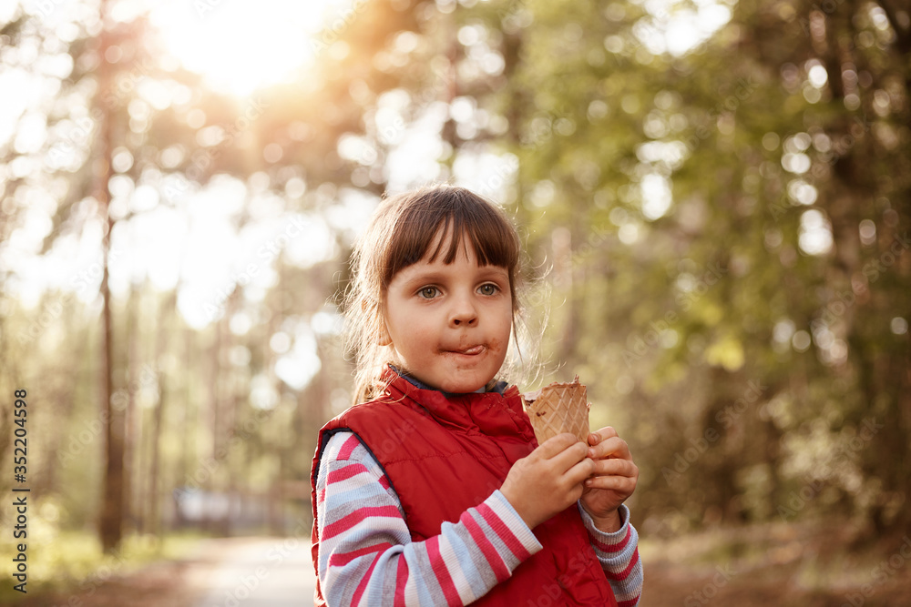 Portrait of pretty playful obedient kid looking ahead, holding ice cream in hands, playing outside, posing over trees background, being in park, having thoughtful facial expression. Rest concept. Portrait of pretty playful obedient kid looking ahead, holding ice cream in hands, playing outside, posing over trees background, being in park, having thoughtful facial expression. Rest concept.