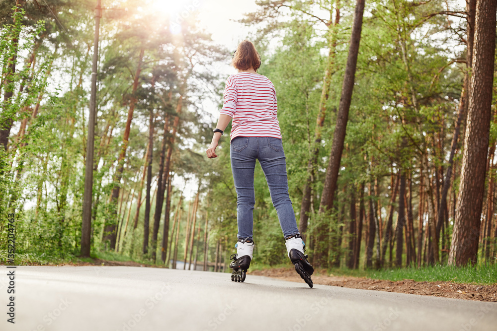 Back view of skilled good looking young sportswoman having hobby, rollerskating, having rest, wearing striped sweatshirt and jeans, having training in forest, spending time in peace. Hobby concept. Back view of skilled good looking young sportswoman having hobby, rollerskating, having rest, wearing striped sweatshirt and jeans, having training in forest, spending time in peace. Hobby concept.
