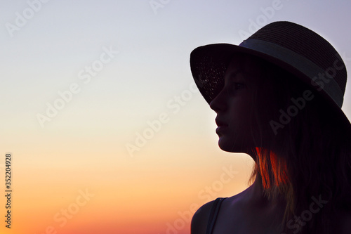 Blurry silhouette of teenage girl wearing a sun hat and looking to the left, close up portait, space for text. Female portrait, sunset background, cropped shot. People, travel concept.