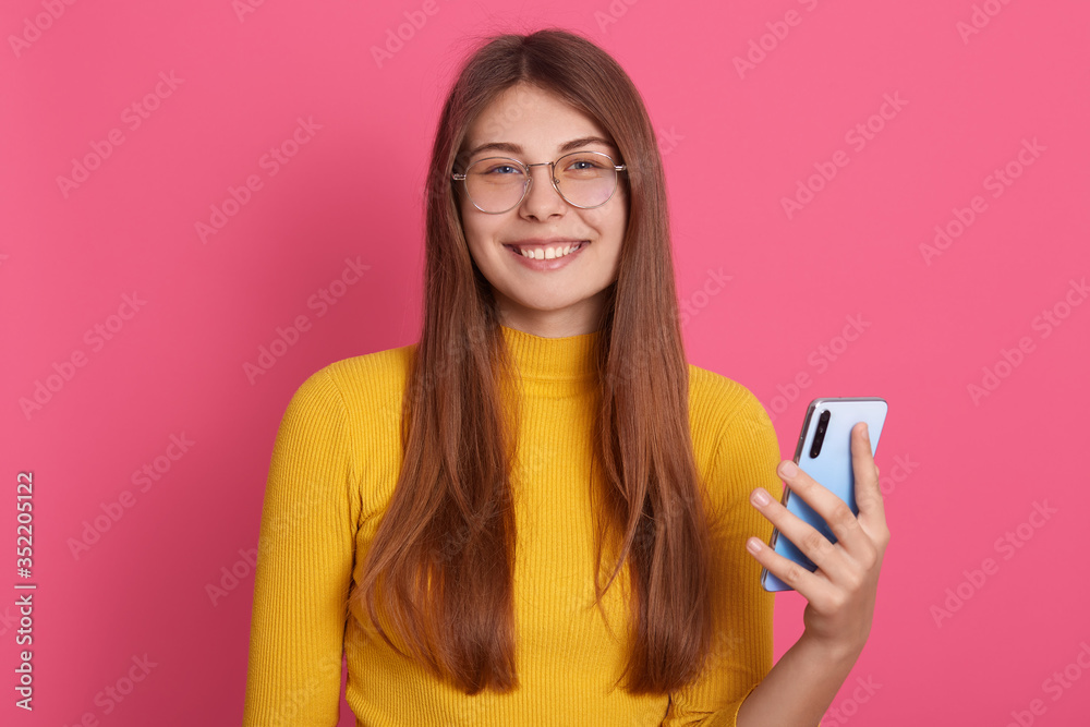 Photo of cheerful positive good looking young female posing isolated over pink background in studio, smiling sincerely, holding smartphone in hand, wearing sweatshirt and spectacles. Tech concept. Photo of cheerful positive good looking young female posing isolated over pink background in studio, smiling sincerely, holding smartphone in hand, wearing sweatshirt and spectacles. Tech concept.