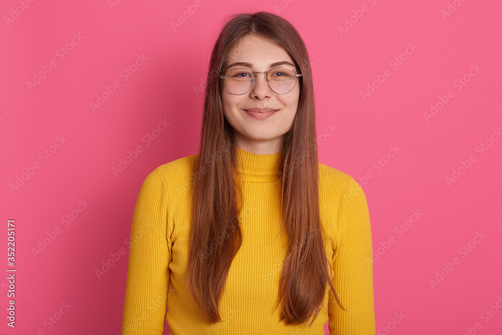 Indoor studio shot of attractive beautiful pretty young woman wearing eyeglasses and yellow sweatshirt, having long fair hair, posing isolated over pink background. People and emotions concept.