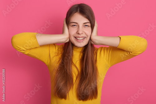 Horizontal picture of cheerful good looking positive young lady having pleasant facial expression, looking directly at camera, being in good mood, wearing casual clothes, covering ears with hands. photo