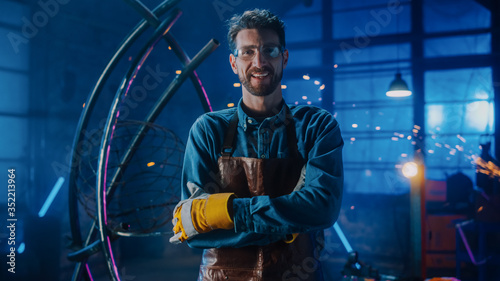 Young Professional Fabricator in Safety Glasses and Apron Gently Smiles at the Camera with Crossed Arms. Authentic Artist Wearing Work Clothes in a Metal Workshop. Sparks Flying on the Background. photo