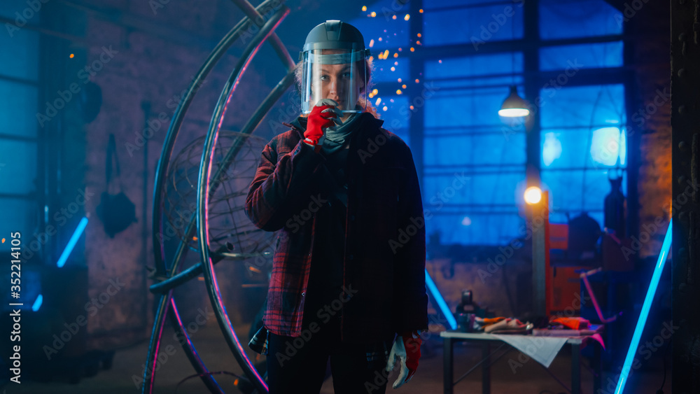 Young Beautiful Empowering Woman with Ear Piercing Wear Safety Mask and Gently Smiles at the Camera. Authentic Fabricator Wearing Work Clothes in a Metal Workshop. Sparks Flying on the Background.