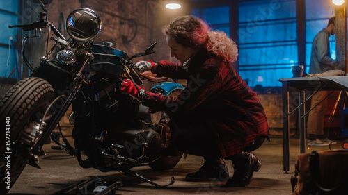 Young Beautiful Female Mechanic is Fixing a Custom Bobber Motorcycle. Talented Girl Wearing a Checkered Shirt. She Uses a Ratchet Spanner. Creative Authentic Workshop Garage.