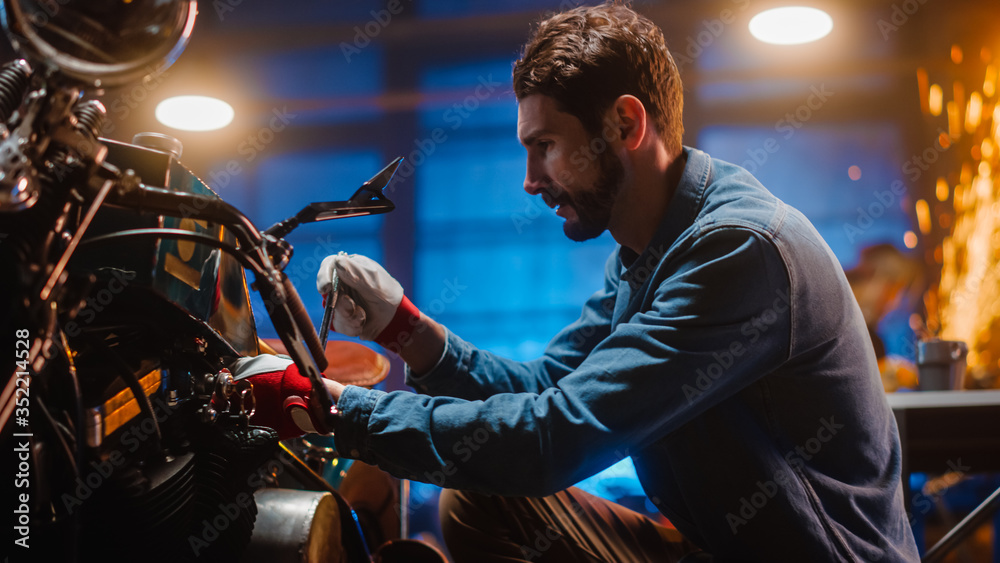 Young Handsome Mechanic is Fixing a Custom Bobber Motorcycle. Close Up on Work Gloves. Talented Man Uses a Ratchet Spanner. Creative Authentic Workshop Garage.