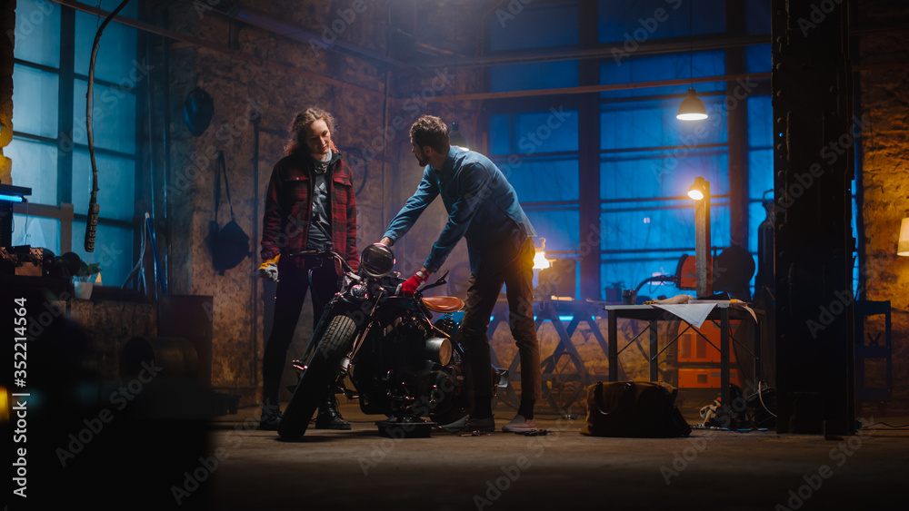 Two Young Professionals are Discussing The Work Done on a Custom Bobber Motorcycle. Hipster Man Explaining the Job to a Talented Female Mechanic. Creative Authentic Workshop Garage.