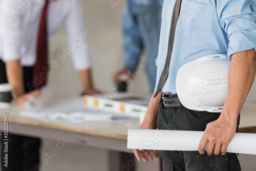 Young engineer holding a hard hat or white helmet and blueprint with worker at the construction site in background - teamwork concepts. photo