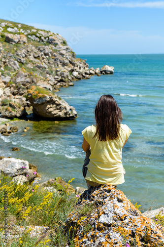 a girl sits with her back to us on a rock on a rocky coast in Sunny weather and looks out to sea