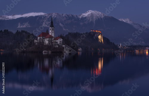 Church Of Mary The Queen, Also Known As The Pilgrimage Church Of Assumption Of Mary, Or Our Lady Of The Lake Bled At Winter « Blue Hour » With Karavanke Mountain Range And Parish Church Of St.Martin