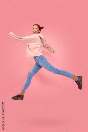 Full length side profile of emotional cheerful beautiful energetic caucasian red-haired female hipster jump on in the air, hurry shopping black Friday, low prices isolated over pink studio background
